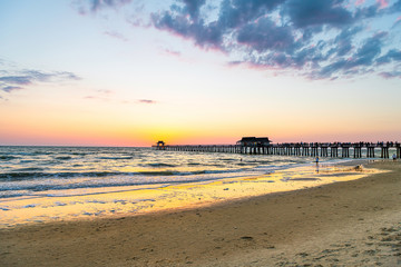 Naples, Florida yellow sunset in gulf of Mexico with sun behind Pier wooden jetty on horizon and silhouette ocean waves wide angle view with sand, people