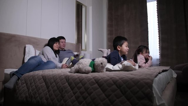 Loving Chinese Parents With Elementary Age Children Spending Joint Domestic Leisure Lying On Bed In Front Of TV-set. Friendly Family Together With Little Kids Watching Movie On TV In Bedroom