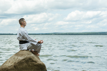 A man in a kimono sits on a rock, under it is the sea. Master with a black belt. Practical class in karate. The technique of meditation. Cleansing and thinking about life before the fight.