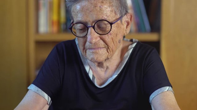 Tilt up of a nice old woman with round glasses using joyfully a smartphone at home living room near a bookcase. Technological ederly lady surfing the internet with a mobile phone. Cine lens.
