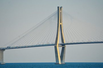 A vertical pylon with radiating steel supports is a feature of a bridge over a bay in South Korea. A bus can be seen on the road of the bridge. The water is blue, and the sky hazy.