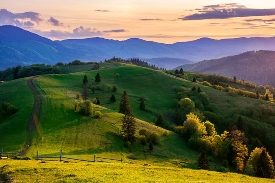 mountainous countryside in springtime at dusk. road, wooden fence and trees on the rolling hills. ridge in the distance. clouds on the sky. beautiful rural landscape of carpathians
