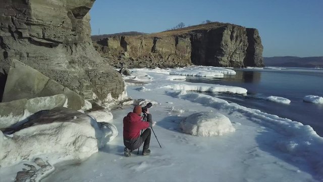 Professional photographer is taking pictures of winter sea, snowy coasline and high cliffs. Beautiful landscape. Sunny day