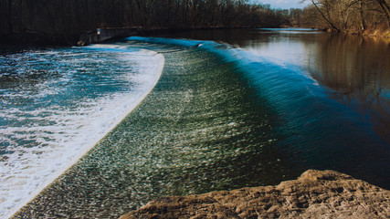 A Fast Flowing Waterfall With Deep Blue Water and White Foam With Trees in the Background and a rock in the foreground