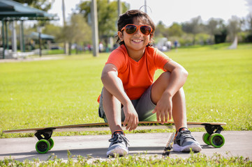 young boy sitting skateboard suntan sidewalk longboard smiling orange glasses shirt green grass park sunny