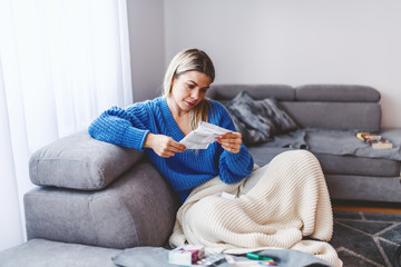 Attractive caucasian blond woman sitting on sofa in living room covered with blanket and reading side effects about new medicine. - obrazy, fototapety, plakaty