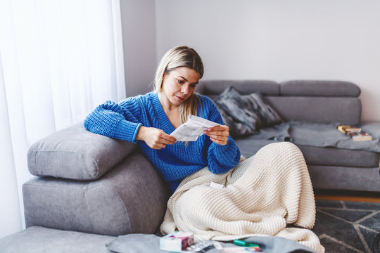 Attractive Caucasian Blond Woman Sitting On Sofa In Living Room Covered With Blanket And Reading Side Effects About New Medicine.