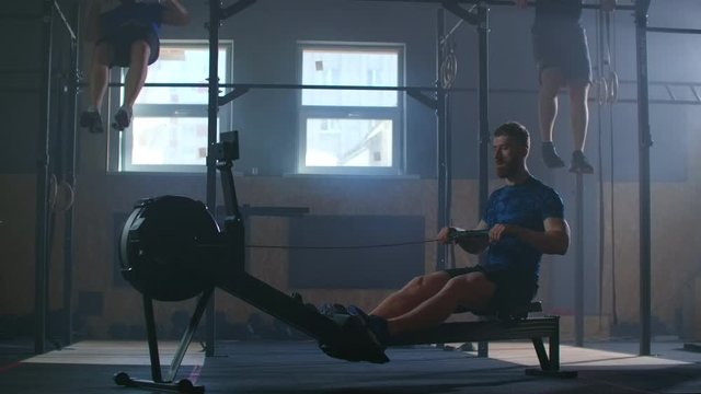 Slow-motion cross-training, three men in the fitness room, two pulling up in the background, one using a rowing machine.
