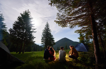 Loving couple and friends at night camping in the mountains. Tourist people sitting by campfire near tents under the spruce trees against the backdrop of the mountain valley.