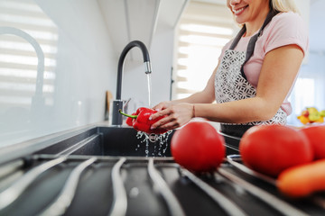 Close up of caucasian worthy woman in apron washing red pepper in kitchen sink. Lunch preparation concept.