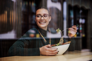 Young smiling college girl sitting in restaurant, having delicious salad for lunch and looking trough window. She is on the break between classes. Picture taken from outside.