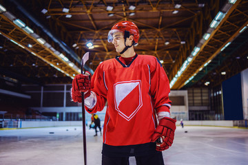 Attractive strong hockey player standing on the ice in hall and holding a stick.
