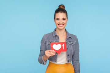 Portrait of joyful beautiful fashionably dressed woman with hair bun holding social media heart icon, network one Like button, follower notification symbol. studio shot isolated on blue background