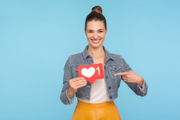 Portrait of cheerful excited fashionably dressed woman with hair bun pointing at social media Heart icon, network one Like button, follower notification symbol. studio shot isolated on blue background