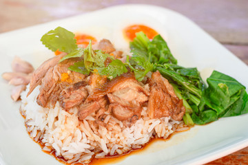 Stewed leg pork (Kao Kha Moo) on rice with Chinese style sweet gravy sauce, eggs, garlic and vegetable in white plate on wooden table. Selective focus.