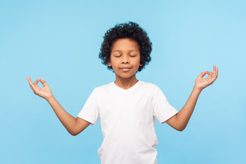 Portrait of peaceful cute little boy holding fingers in mudra gesture and meditating with closed...