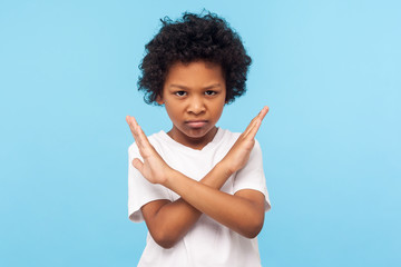 Stop, I'm warning. Portrait of angry determined little boy with curls crossing hands and looking at camera with aggression, showing stop gesture, way prohibited. indoor studio shot blue background