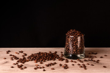 Whiskey glass full of roasted coffee beans and some grains scattered on a wooden surface