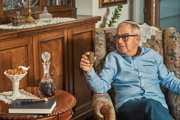 Senior Jew in armchair drinks wine in traditional home 