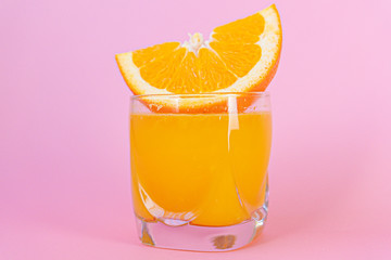 Orange juice and piece of orange with pink background.