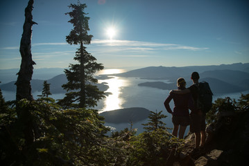 COUPLE STANDING on the top of mountain overlooking islands during sunset in beautiful British Columbia Vancouver Canada after hike in forest