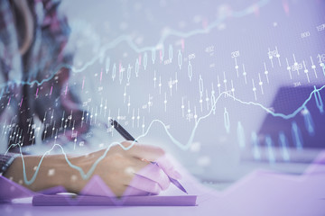 A woman hands writing information about stock market in notepad. Forex chart holograms in front. Concept of research. Double exposure