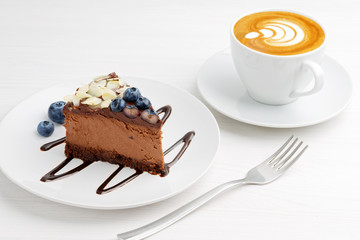 Closeup cup of coffee cappuccino and piece of homemade chocolate cheesecake with blueberries and almond slices on white wooden table. Angle view.
