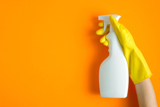 Hand in yellow rubber glove holding plastic spray bottle with cleaning detergent on a orange background