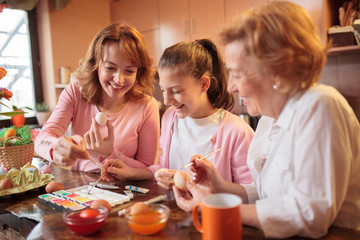 Teenage caucasian girl painting and decorating Easter eggs with her mother and grandmother in home kitchen
