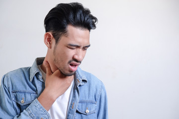 Fototapeta premium Young man have a sore throat and touching his neck, wearing white t-shirt and blue jeans jacket against light grey background, hard to swallow, wuhan virus infection and public widespread