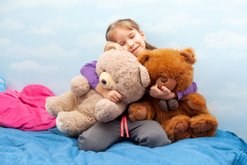 Little girl, smiling child sitting on the bed hugging two big teddy bears, cheerful kid holding her two toys close. Blue room clouds background, heart shaped pillow Love, childhood cuddly toys concept
