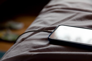 A simple modern no name smartphone laying in bed in crumpled white sheets charging in the morning,...