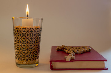 Lighted candle standing in transparent glass in dried peas, nearby lying small prayer book with hard pink binding with simple wooden Christian Catholic rosary on it.  Concept: Simple daily prayer for 