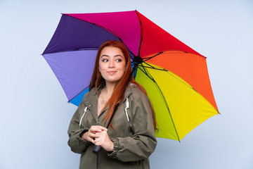 Redhead teenager girl holding an umbrella over isolated blue background laughing