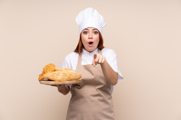 Redhead teenager girl in chef uniform. Female baker holding a table with several breads surprised and pointing front