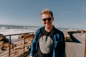 Fototapeta premium Smiling happy european man with blond hair enjoying his trip and walking near the ocean in sunny day