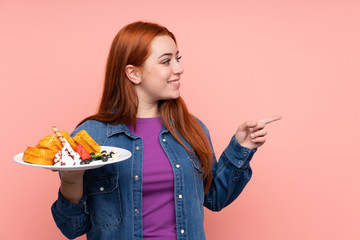 Redhead teenager girl holding waffles over isolated pink background pointing to the side to present a product