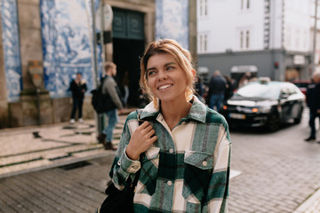 Fototapeta premium Close-up portrait of cheerful white woman with backpack on blur background. Photo of fashionable girl with beautiful brown hair smiling to camera.