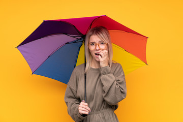 Young blonde woman holding an umbrella over isolated yellow wall nervous and scared