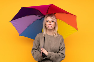 Young blonde woman holding an umbrella over isolated yellow wall pleading