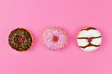 Three donuts with pink, white and brown chocolate glazing and sugar sprinkles in a row on pink background