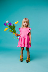 little blonde girl in big yellow shoes and a pink dress is holding spring flowers, baby girl in a pink dress and mother's shoes smiling on a blue background, a little fashionista. space for text