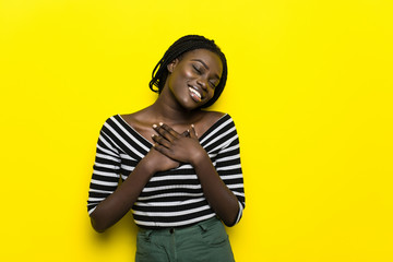 Headshot of pleased touched young African American woman smiles gently, feels pleased, wears striped top and headwear, isolated over yellow background. People, happiness and emotions concept