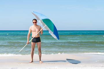 Fit muscular man on ocean beach shore of Santa Rosa, Panhandle Florida with sunglasses holding umbrella with waves crashing in background