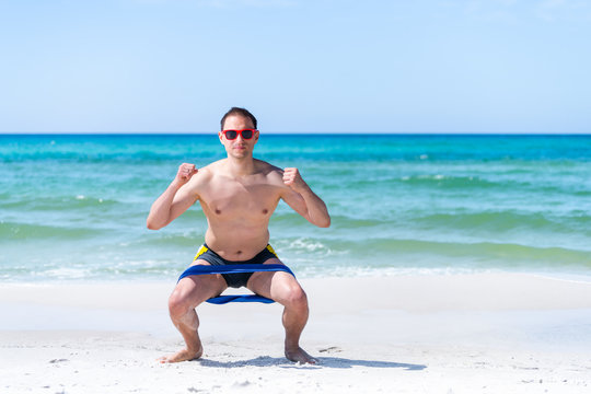 Young Adult Man Squatting At Beach Working Out With Elastic Resistance Band For Legs Exercise With Ocean Background