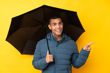 Young handsome man holding an umbrella over isolated yellow background surprised and pointing...