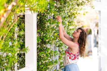 Young adult woman in summer on sunny day in garden touching vine plant flowers outside gardening...