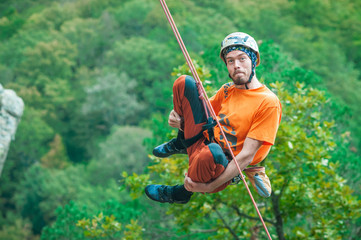 A funny, naughty climber in an orange shirt swinging on a rope on the background of green forest,...