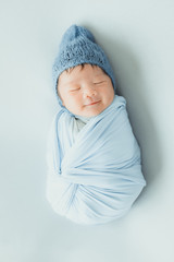 An adorable cute smiling Asian newborn baby with blue knitted hat and wrapprd in cocoon sleeping on bed