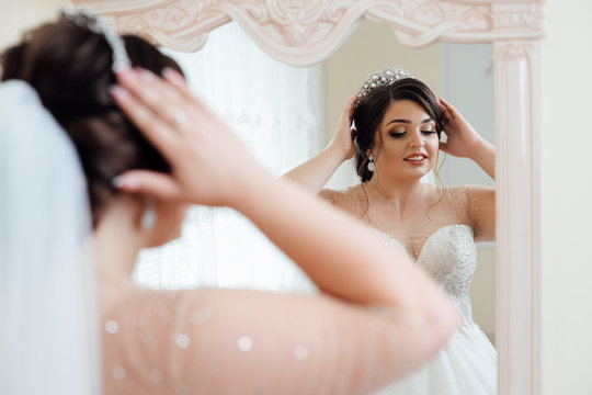 Young cute bride with beautiful hairdo morning at home in dress. Wedding style. Beautiful young bride with luxury wedding hairstyle.The bride looks in the mirror.